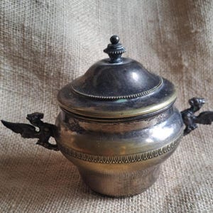 Puede incluir: Azucarero antiguo plateado con tapa y dos asas decorativas con forma de criaturas aladas. El cuenco tiene un diseño detallado alrededor de la base y un pequeño adorno en la tapa. El metal tiene un aspecto deslustrado.