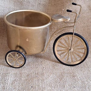 Op de afbeelding: Een decoratieve messing driewieler plantenbak met een grote emmer-vormige voorkant en twee kleinere wielen. De wielen hebben zwarte rubberen banden en gouden spaken. De driewieler heeft een klein zadel en een stuur.