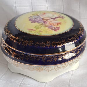Könnte beinhalten: Runde Schmuckdose aus Porzellan mit marineblauem Äußeren und goldenen floralen Mustern. Der Deckel hat eine gelbe Mitte mit einem Blumenstrauß. Die weiße Basis ist mit goldenen Details verziert.