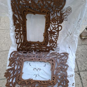 Op de afbeelding: Twee bruine houten fotolijsten met ingewikkelde, opengewerkte houtsnijwerken van bloem- en bladmotieven. De lijsten hebben een rechthoekige centrale opening en een gedetailleerd, organisch randontwerp met zichtbare houtnerf.