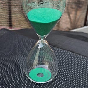 Könnte beinhalten: Eine transparente Glas-Sanduhr mit grünem Sand. Die Sanduhr hat eine bauchige Ober- und Unterseite, die durch einen schmalen Hals verbunden sind. Der grüne Sand ist im oberen Teil sichtbar. Die Sanduhr steht auf einer dunklen Oberfläche.