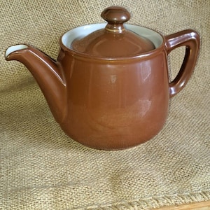 Può includere: Una teiera in ceramica marrone con coperchio e manico abbinati. La teiera ha un interno bianco e un beccuccio curvo. La teiera è appoggiata su una superficie di juta.