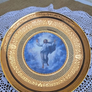 Peut inclure: Une assiette en porcelaine avec un bord doré, un fond noir et un motif doré. Le centre de l'assiette présente une image bleue et blanche de Jésus montant au ciel.