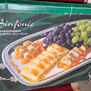 Può includere: Un vassoio da portata argentato con manici decorati, che presenta un assortimento di uva, formaggio e altri spuntini. Il vassoio è etichettato "Sinfonie Sandwichtablett mit Griffen 54,5 x 32 cm".