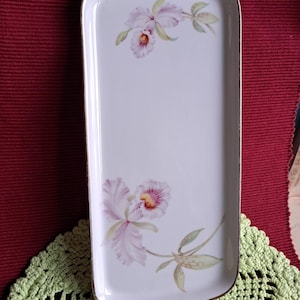 Op de afbeelding: Rechthoekige witte serveerschaal met gouden rand en handvatten. De schaal is versierd met een bloemenontwerp met roze en paarse orchideeën en groene bladeren. De schaal is waarschijnlijk van porselein.
