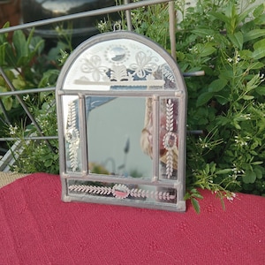 Peut inclure: Un petit miroir en forme d'arche avec un cadre argenté. Le miroir présente des motifs floraux et de feuilles gravés. Le cadre a une forme rectangulaire avec un sommet en arc. Le miroir est posé sur une surface rouge, entouré de verdure.