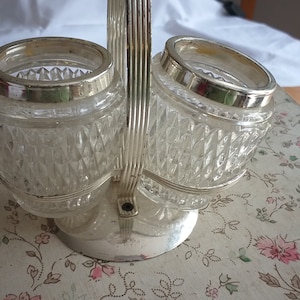 Op de afbeelding: Een zilverkleurige dubbele pottenhouder met twee heldere glazen potten met een diamantpatroon. De potten staan op een wit oppervlak met een bloemenpatroon.
