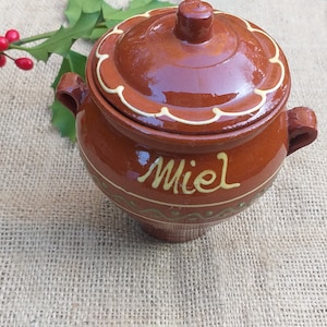 Könnte beinhalten: Ein brauner Keramik-Honigtopf mit Deckel und einer weißen Aufschrift "Miel". Der Topf ist mit einem weißen gewellten Rand und einem weißen Blumenmuster verziert.