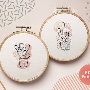 Op de afbeelding: Twee borduurringen met kunstwerken met cactussen. De cactus links is een vijg cactus met roze en bruine accenten. De cactus rechts is een hoge, slanke cactus met gele en bruine accenten. Beide ringen hebben een witte achtergrond en een houten frame.