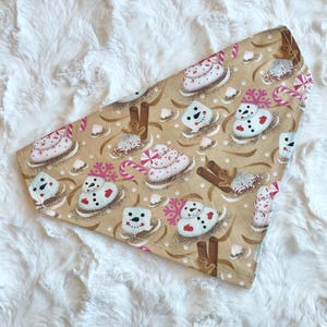May include: A brown bandana with a white background and a repeating pattern of snowmen, candy canes, and gingerbread men. The bandana is trimmed with a white border.