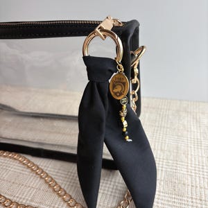 May include: A clear plastic bag with black trim and a gold chain strap. A black fabric bow is attached to the gold hardware, featuring a gold oval charm with a helmet graphic and the year 1985, followed by small lettered beads spelling RM.