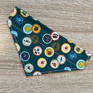 May include: A green bandana with a white stitched border features a pattern of colorful, circular patches with camping and outdoor-themed illustrations and text. The patches include phrases like "Go Outdoors", "Happy Camper", "Little Explorer", and "Let's Go For A Hike".