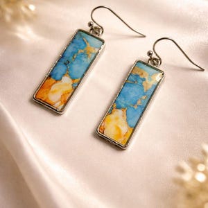 May include: Two rectangular earrings with silver-coloured frames. Each earring has a blue, yellow, and orange stone with a marbled pattern. The earrings are presented on a white, satin-like surface.
