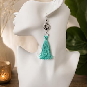 Può includere: Un orecchino pendente caratterizzato da un ciondolo circolare color argento con un intricato motivo a filigrana traforata, collegato a una nappa in seta color turchese brillante. L'orecchino pende da un gancio color argento standard e sottile.