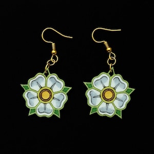 May include: A pair of earrings featuring a white flower design with a yellow centre and green leaves. The earrings have gold-coloured hooks. The background is black.