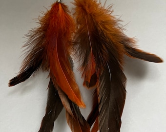 May include: A pair of feather earrings with silver-colored hooks. The earrings feature a combination of brown, orange, and black feathers. The feathers are arranged in a layered design, creating a full and textured appearance. The earrings are designed for pierced ears.