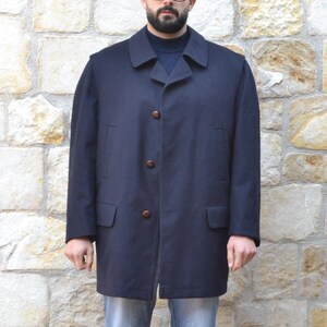 Può includere: Un cappotto blu navy con bottoni, indossato sopra un maglione blu scuro e jeans blu chiaro. La persona indossa scarpe da ginnastica verdi e bianche. Lo sfondo è un muro di pietra.