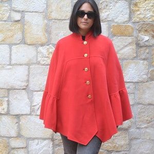 May include: A woman wearing a red cape with gold buttons and black sunglasses. She is standing in front of a stone wall and wearing grey jeans and black shoes.