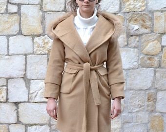 May include: A camel-colored wrap coat with a fur-trimmed hood, a belt, and two pockets. The coat is worn over a white turtleneck sweater and black pants. The person is wearing white sneakers.