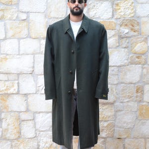 May include: A long, dark green overcoat with a traditional design. The coat has a notched collar, button fastening, and a tailored fit. The model is wearing a white t-shirt, light blue jeans, and grey trainers.