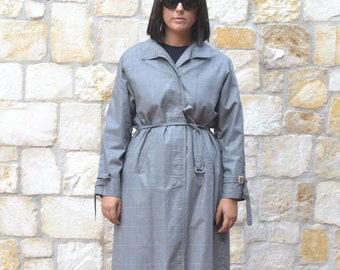 Könnte beinhalten: Ein grauer karierter Trenchcoat mit Gürtel und schwarzer Sonnenbrille. Der Mantel wird von einer Person getragen, die vor einer Steinmauer steht.