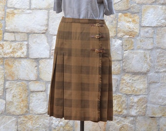 May include: A brown and tan plaid skirt with a fringed hem and three gold buckles. The skirt is on a mannequin wearing a light gray sweater.