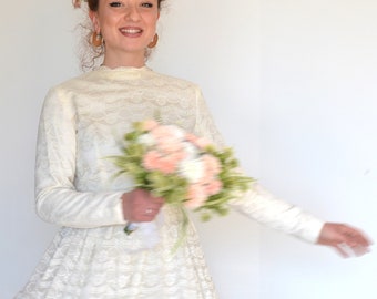 May include: A woman in a white lace dress with long sleeves and a full skirt. She is wearing a flower crown and holding a bouquet of pink and white flowers.