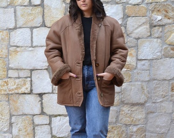 May include: A brown shearling jacket with a fur collar and pockets. The jacket is worn over a black turtleneck and blue jeans. The person is wearing black shoes and sunglasses.