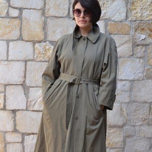 May include: A woman wearing a long, olive green trench coat with a belt and a button closure. She is wearing sunglasses and standing in front of a stone wall.