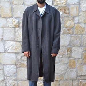 May include: A long, dark gray wool coat with a button closure. The coat has a relaxed fit and is worn over a white t-shirt and blue jeans. The person is wearing brown leather boots.