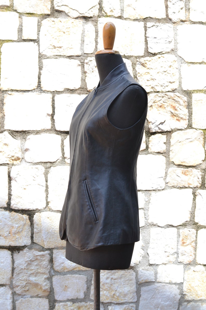 May include: A black leather vest with a zipper closure and a pocket on the front. The vest is on a mannequin against a white stone wall.
