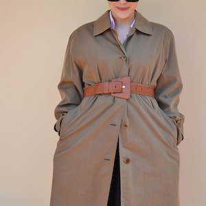 Könnte beinhalten: Eine Frau trägt einen braunen Trenchcoat mit einem braunen Ledergürtel. Sie trägt eine schwarze Sonnenbrille und hat kurze braune Haare.