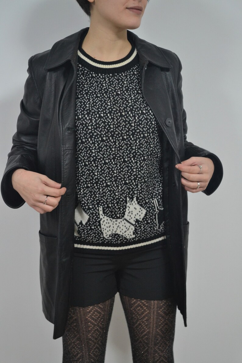 May include: A black leather jacket with a black and white patterned sweater featuring a white dog design. The person is wearing black shorts and black patterned tights.