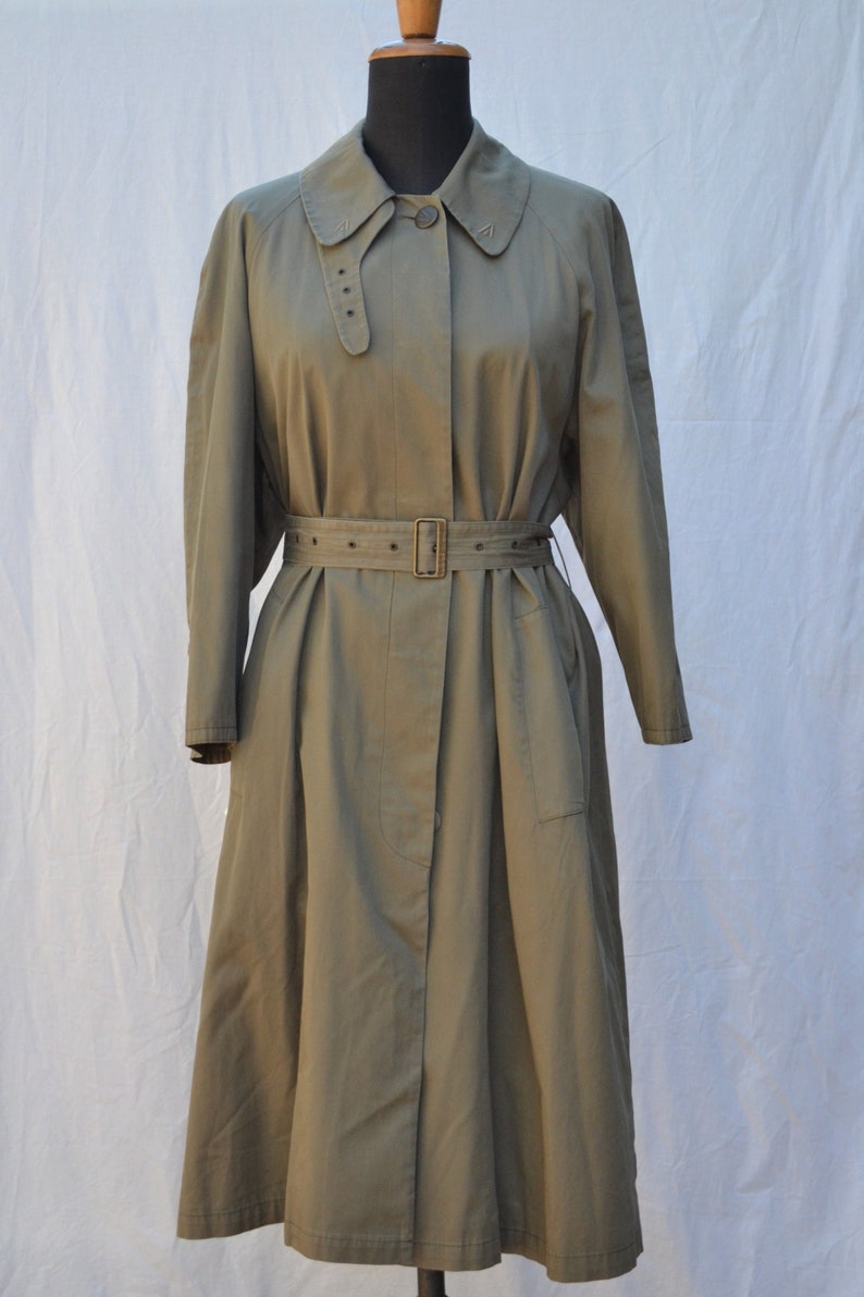 May include: A long, olive green trench coat with a belt and a button closure. The coat has a collar and long sleeves.