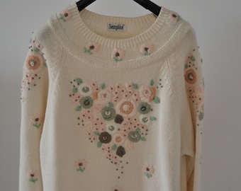 May include: A cream knitted jumper with a floral embroidery design. The jumper has a crew neck and long sleeves. The embroidery features a heart-shaped bouquet of pink, peach, and green flowers.