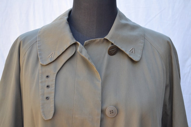 May include: A green, long-sleeved jacket with a collar and two buttons. The jacket has a small logo on the collar and a larger logo on the button.