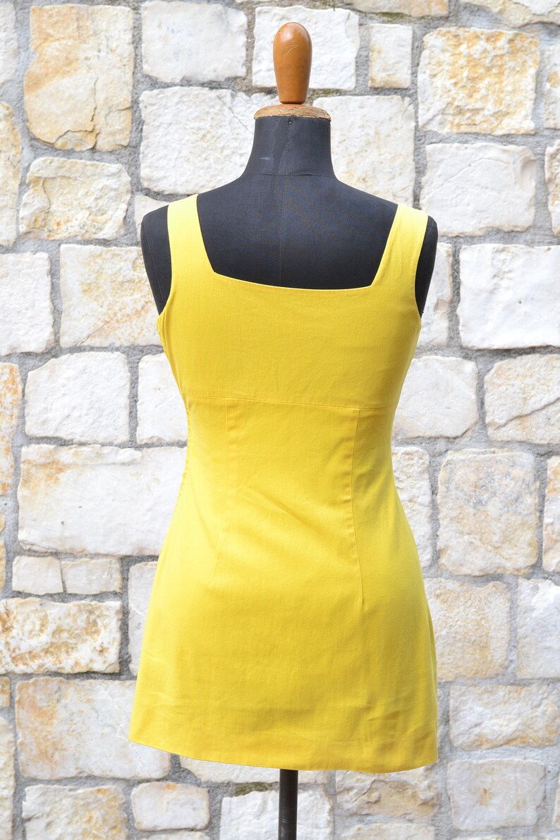 May include: A sleeveless yellow dress with a square neckline. The dress is fitted and has a straight skirt.