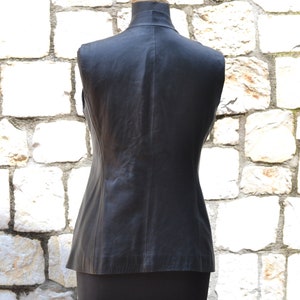 May include: A black leather vest with a sleeveless design. The vest is on a mannequin against a white stone wall.
