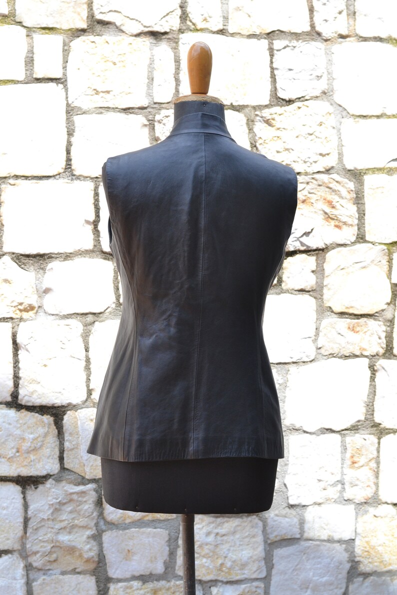 May include: A black leather vest with a sleeveless design. The vest is on a mannequin against a white stone wall.