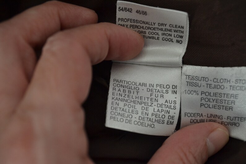 K&ouml;nnte beinhalten: Nahaufnahme eines braunen Stofflabels mit wei&szlig;em Text. Der Text lautet "Professionally dry clean only, perchloroethylene with colors cool iron low, tumble cool no." Das Etikett enth&auml;lt auch Details zur Stoffzusammensetzung, darunter "100% poliestere, polyester, poliester" und "Fodera-lining-futter, doublure, forro".