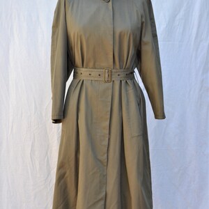 May include: A long, olive green trench coat with a belted waist and a high collar. The coat has a single button closure and a flap pocket on the left side.