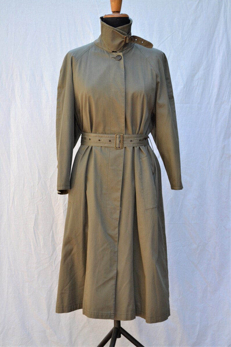 May include: A long, olive green trench coat with a belted waist and a high collar. The coat has a single button closure and a flap pocket on the left side.