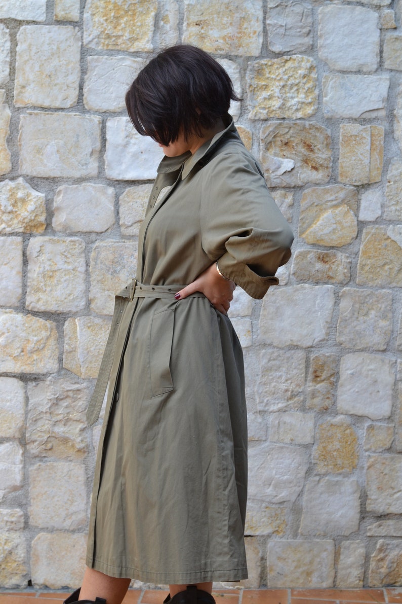 May include: A khaki trench coat with a belt and a button closure. The coat has a long sleeve and a relaxed fit.