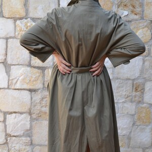 May include: A long, olive green trench coat with a belt. The coat has a single-breasted closure and a long, flowing skirt.
