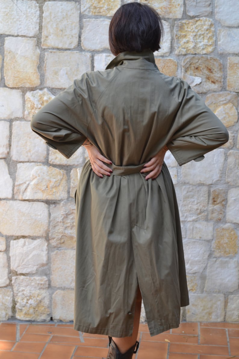 May include: A long, olive green trench coat with a belt. The coat has a single-breasted closure and a long, flowing skirt.