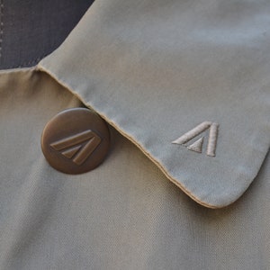 May include: A beige trench coat with a brown button featuring a stylized triangle logo. The collar of the coat is folded over and has a stitched triangle logo.