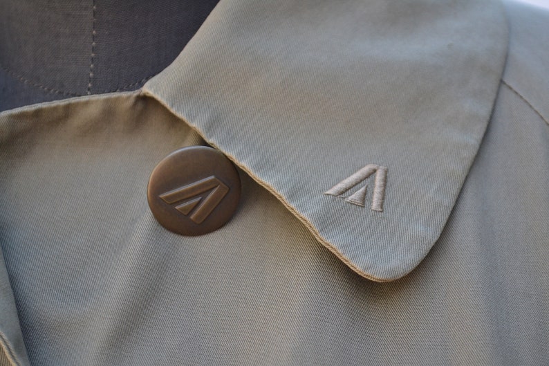 May include: A beige trench coat with a brown button featuring a stylized triangle logo. The collar of the coat is folded over and has a stitched triangle logo.