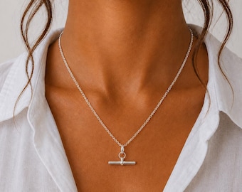 May include: A silver-toned necklace featuring a delicate chain and a horizontal bar pendant attached to a small circular ring. The metal has a polished, reflective finish, and the necklace rests against the collarbone area of a white collared shirt.