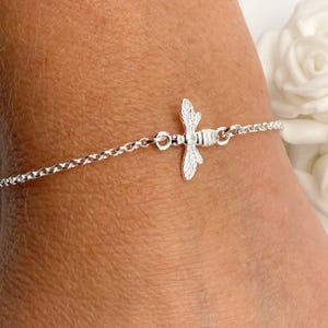 Peut inclure: Un bracelet argenté orné d'un charm en forme d'abeille. L'abeille est le point central, avec des détails complexes sur les ailes et le corps. La chaîne du bracelet est délicate, avec de petits maillons. Le bracelet est porté sur un poignet.