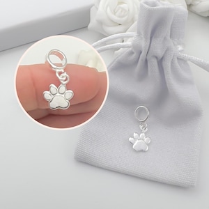 May include: A silver paw print charm with a small ring for attachment. The charm is displayed next to a white drawstring bag and a close-up of the charm held by a person.